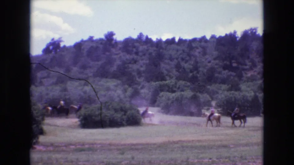 Horseback riders pass through a dusty section of a grassy Colorado field, with bushes, a wooded rise, and soft 8mm color visible.