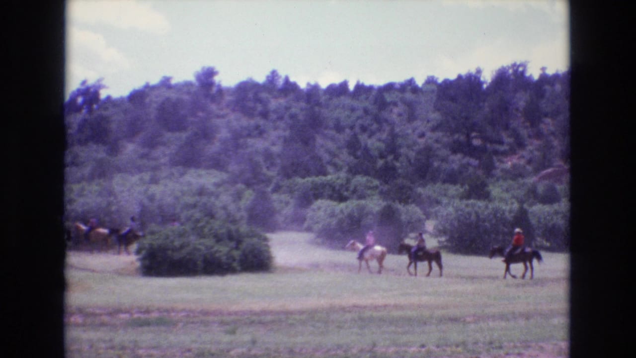 In 1973 Colorado home-movie footage, several people ride horses across an open grassy field below a wooded hillside.