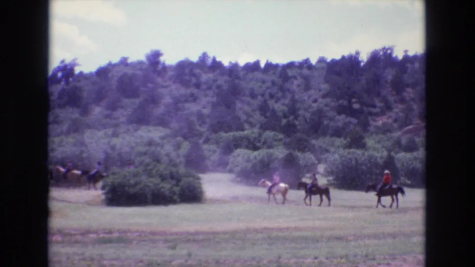 In 1973 Colorado home-movie footage, several people ride horses across an open grassy field below a wooded hillside.