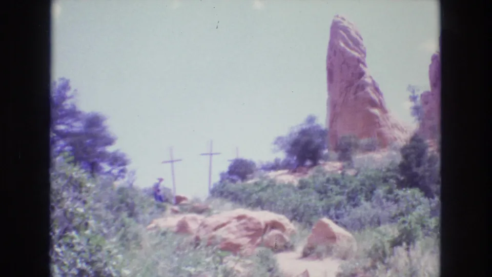 Visitors walk near shrubs and boulders below a red rock formation with three wooden crosses visible on the hillside in Colorado footage from 1973.