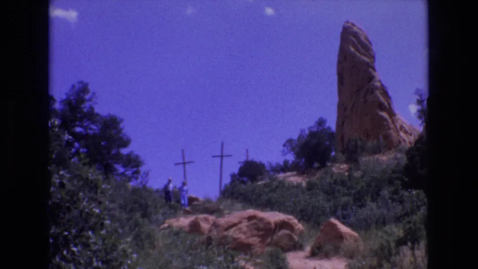 A low-angle 1973 Colorado 8mm frame shows visitors below a red rock formation, with wooden crosses, trees, boulders, and deep blue sky.