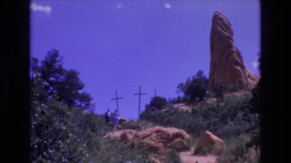 The final 1973 Colorado frame shows small visitor figures on a hillside below wooden crosses and a red rock formation, framed by shrubs and blue sky.