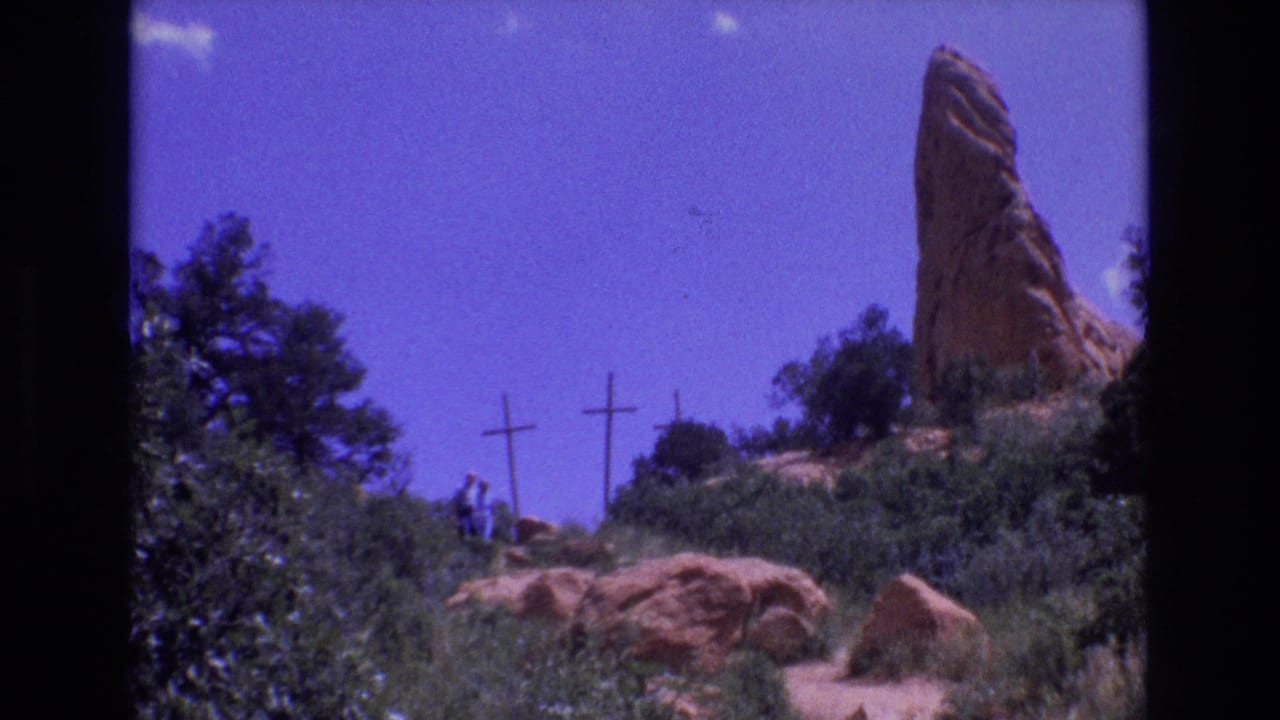 Distant walkers near wooden crosses and a tall red rock spire on a scrubby hillside.