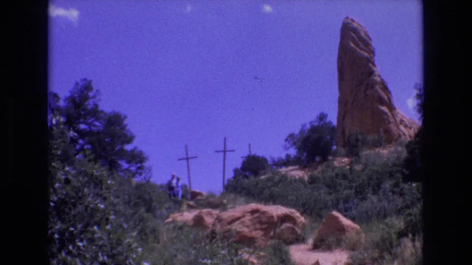 1973 Colorado 8mm frame showing distant walkers on a rocky path below three wooden crosses and a tall red sandstone spire under blue sky.
