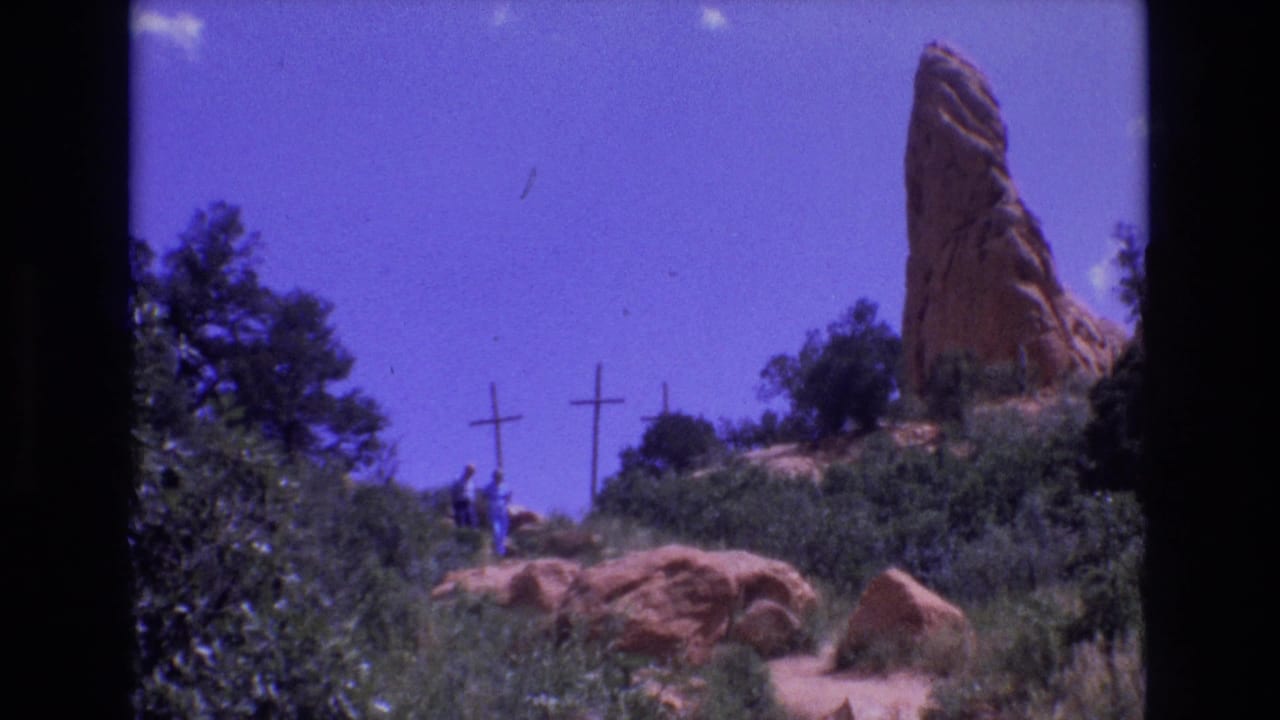 Two distant people stand or walk near crosses with a red rock spire at right.