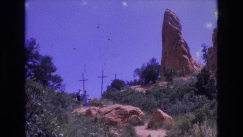 Soft 1973 Colorado 8mm frame showing distant walkers below three wooden crosses, with a red sandstone spire, shrubs, trees, and reddish boulders around the trail.