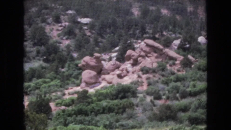 1973 Colorado 8mm frame looking across a scrub-covered rocky hillside with clusters of boulders, trees, and a partial red rock formation along the right edge.