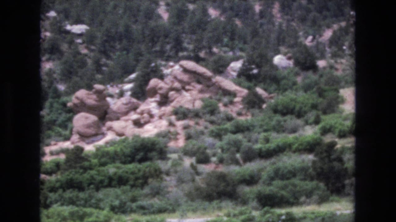 1973 Colorado 8mm frame showing red rock outcrops, pine-covered slopes, and a partly visible road below from a distant handheld viewpoint.