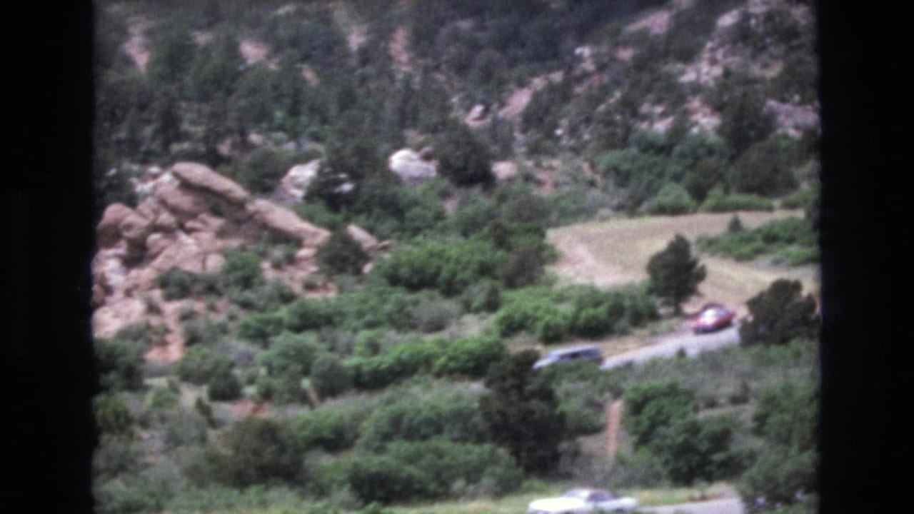 Wooded mountain slope with red rocks and small cars on a road below.