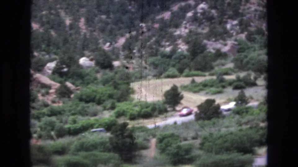 1973 Colorado 8mm frame showing several distant cars on a mountain road beneath red rock outcrops, trees, scrub, and pale open ground.