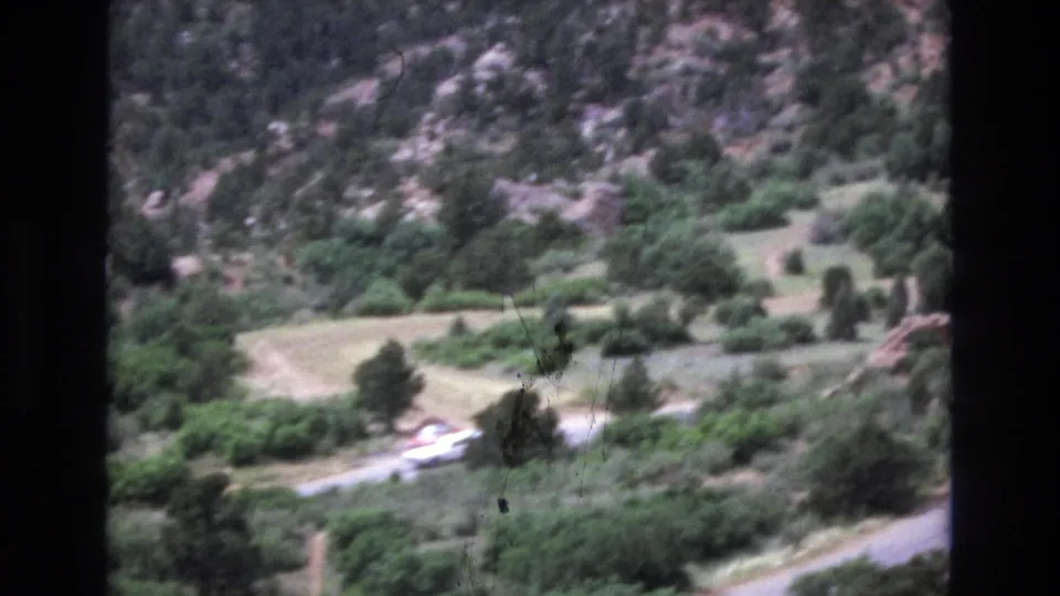 1973 Colorado 8mm frame with a curved road, distant moving cars, wooded slopes, scrub vegetation, and red rock terrain seen through soft handheld footage.