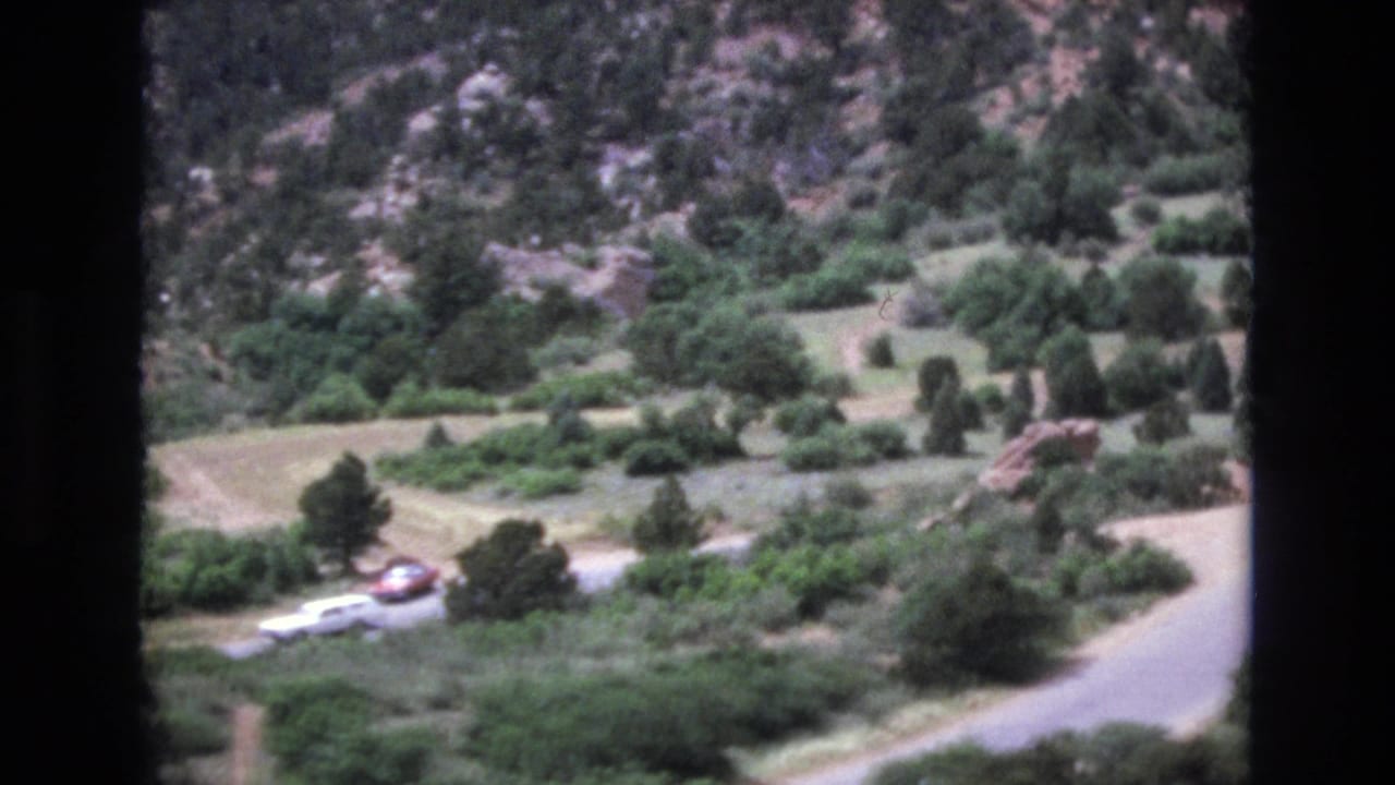 Winding mountain road with distant cars, shrubs, trees, and rocky hillside.