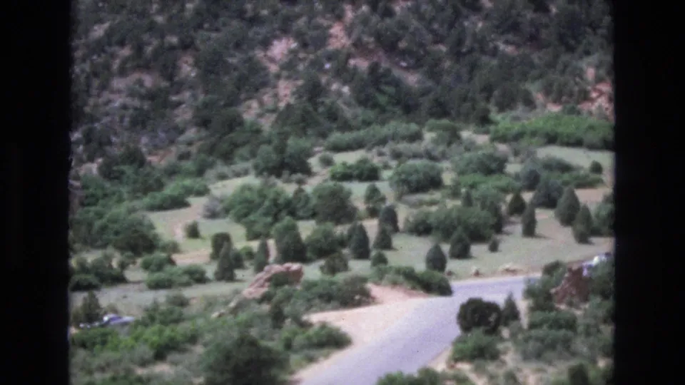1973 Colorado 8mm frame looking down on a paved road through a green valley, with shrubs, evergreens, open grass, a rocky hillside, and a partially visible distant car.