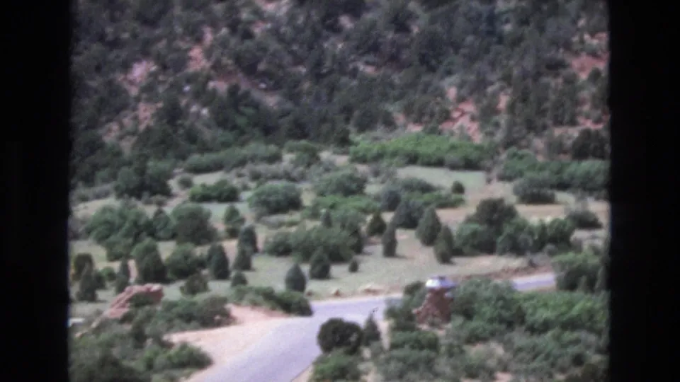 1973 Colorado 8mm frame showing a curving valley road at the base of wooded hills, with a distant car, dense shrubs, grass, evergreens, and reddish rock formations.