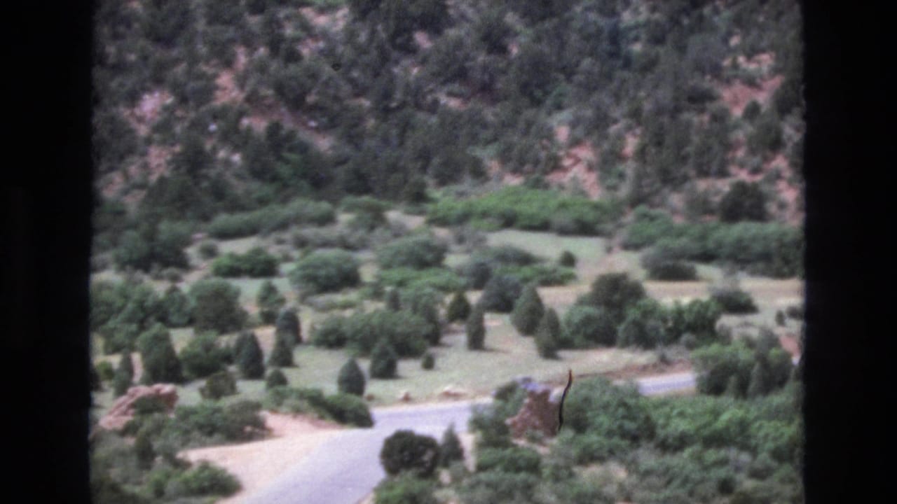 1973 Colorado home-movie frame looking over a mountain valley with a winding road, open ground, scattered evergreens, and a rocky hillside beyond.