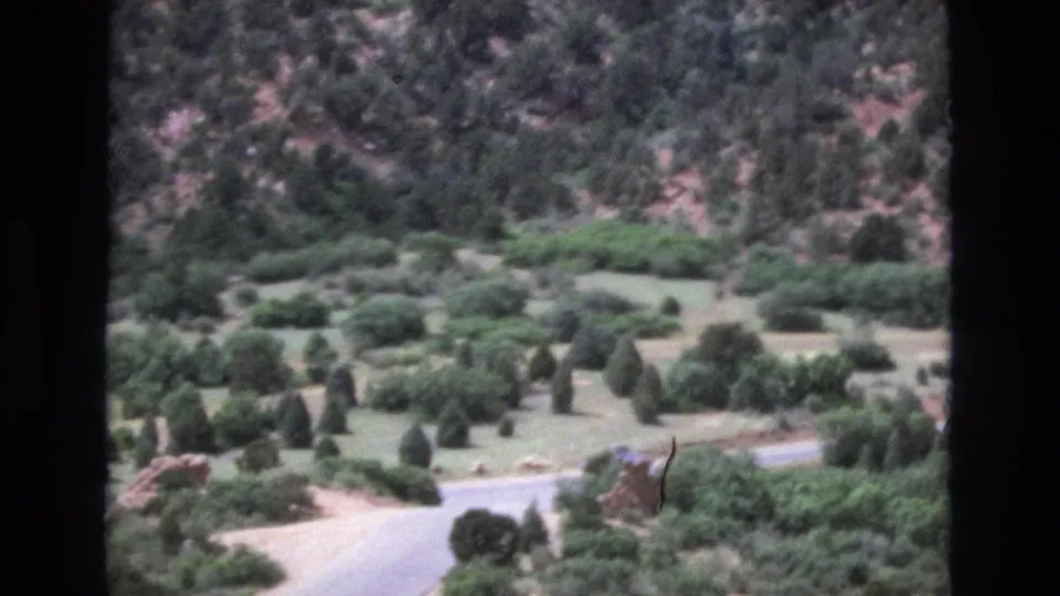 1973 Colorado home-movie frame looking over a mountain valley with a winding road, open ground, scattered evergreens, and a rocky hillside beyond.