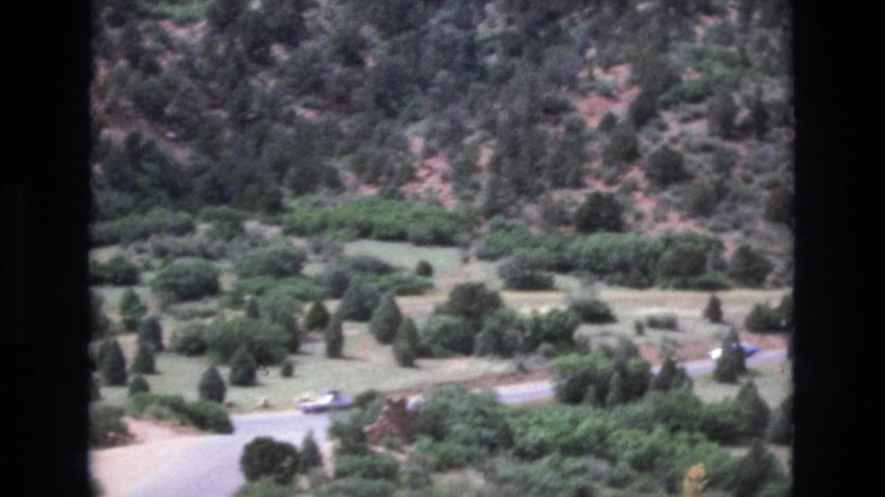 Valley road curving through evergreens with a small light-colored vehicle visible.