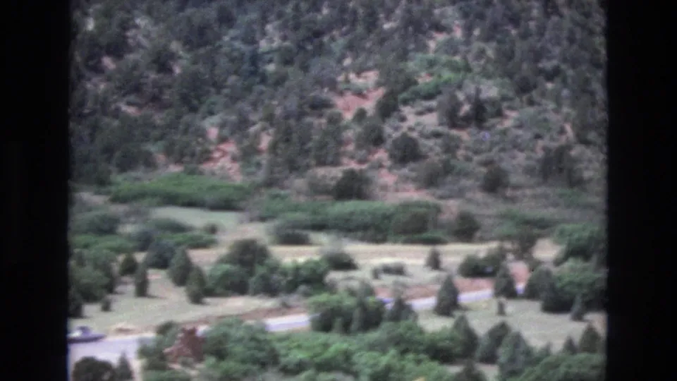 1973 Colorado 8mm frame showing a closer view of a tree-dotted valley, brushy hillside, and a road crossing near the bottom of the frame.