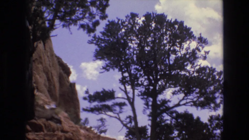 1973 Colorado 8mm frame showing a dark evergreen tree silhouetted beside the edge of a red sandstone wall, with broken clouds across the sky.