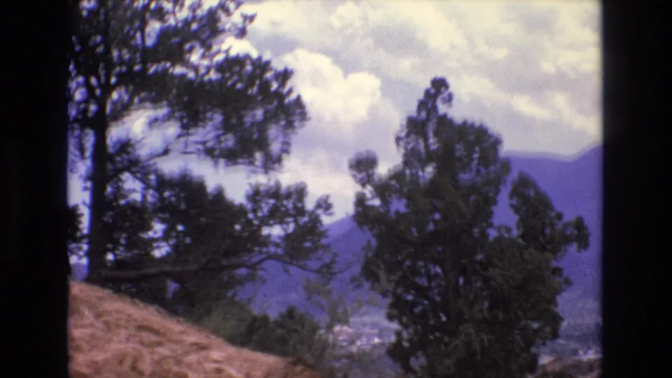 1973 Colorado 8mm frame looking past hillside evergreens toward a hazy mountain valley under broken clouds.
