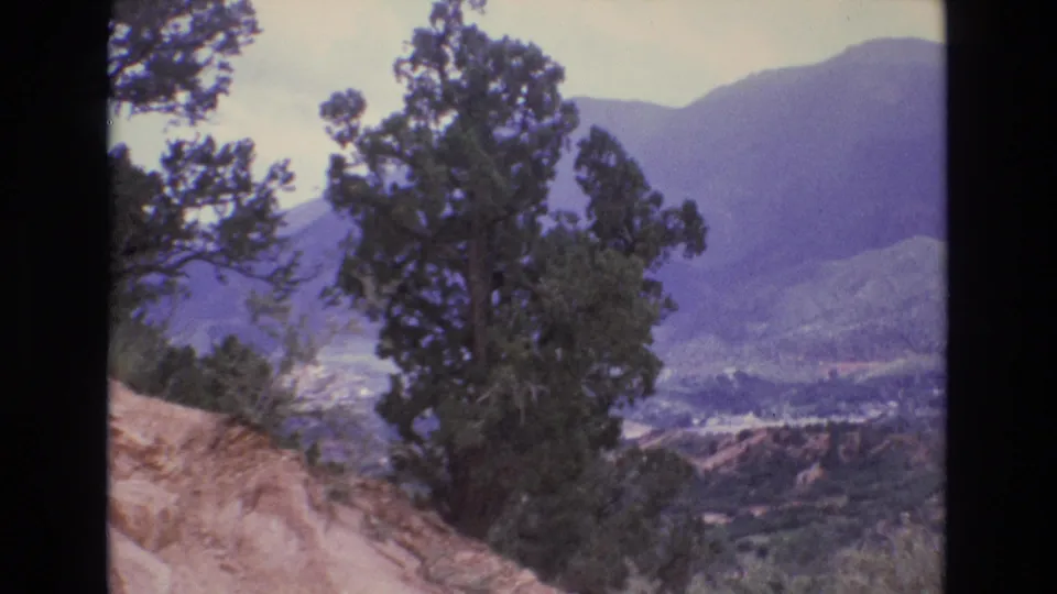 1973 Colorado 8mm frame ending on a hillside evergreen with layered mountains and a sunlit valley visible in the distance.