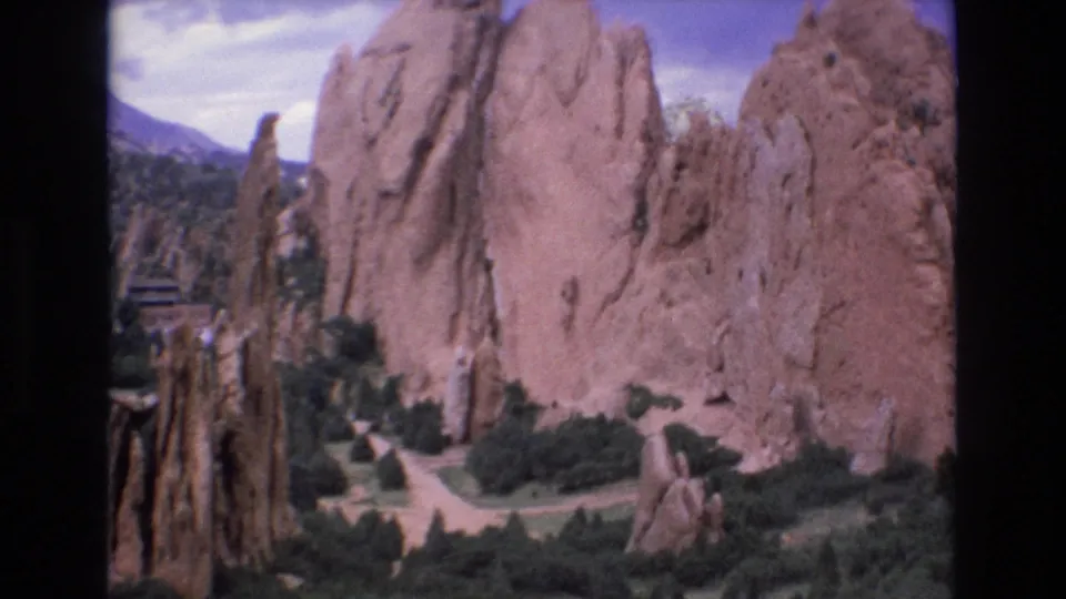 1973 Colorado home-movie frame with a broad red sandstone wall, smaller rock formations, winding paths, and green vegetation below.