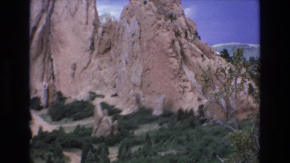 1973 Colorado 8mm frame of Garden of the Gods with a sandstone cliff face, winding path, dense shrubs, bare branches, and sky in a soft home-movie scan.