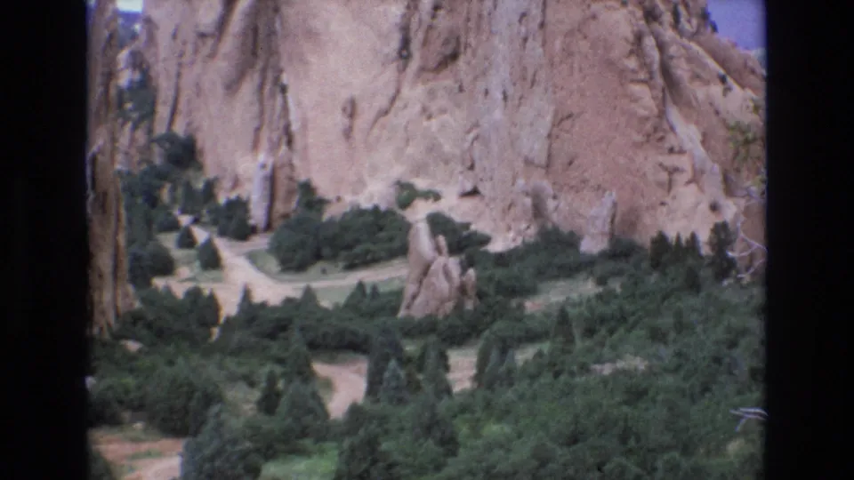 1973 Colorado home-movie frame with a rock spire in the foreground, pale paths crossing green vegetation, and tall sandstone formations behind it.