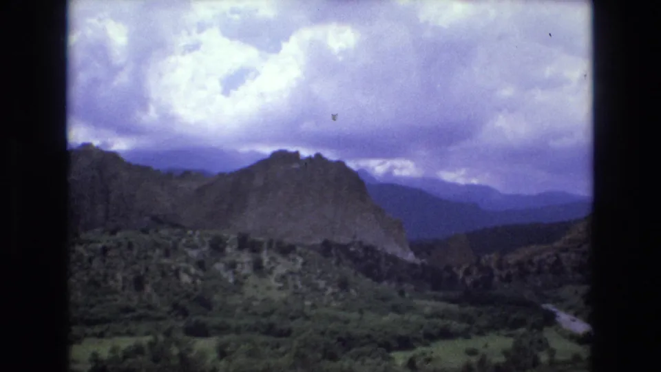 1973 Colorado home-movie frame showing a dark mountain ridge, green forested slopes, distant hills, and a dramatic cloud layer across the sky.