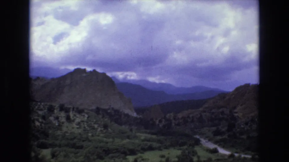 1973 Colorado 8mm frame showing rocky canyon terrain, green valley vegetation, layered distant mountains, and a winding road beneath heavy clouds.