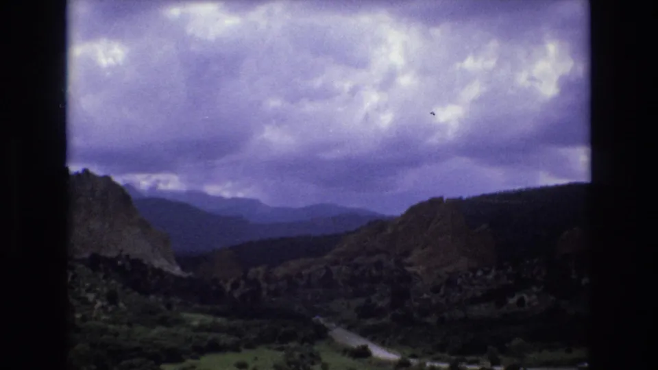 1973 Colorado 8mm frame showing the pan continuing across a canyon valley with a partial rocky wall, winding road, green vegetation, and heavy cloud cover.