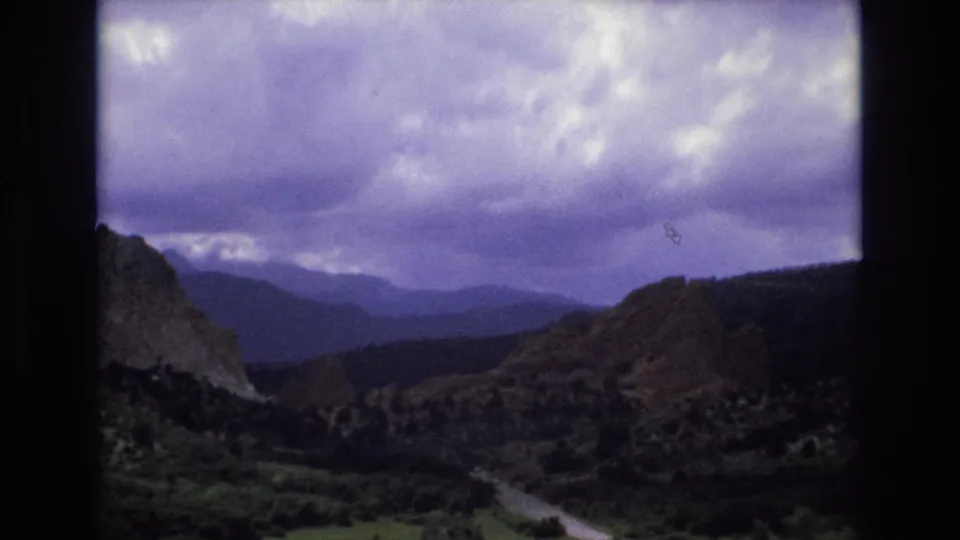 1973 Colorado 8mm frame showing layered mountain ridges, a canyon road, dark rocky slopes, green valley growth, and purple-blue overcast sky from aged color film.