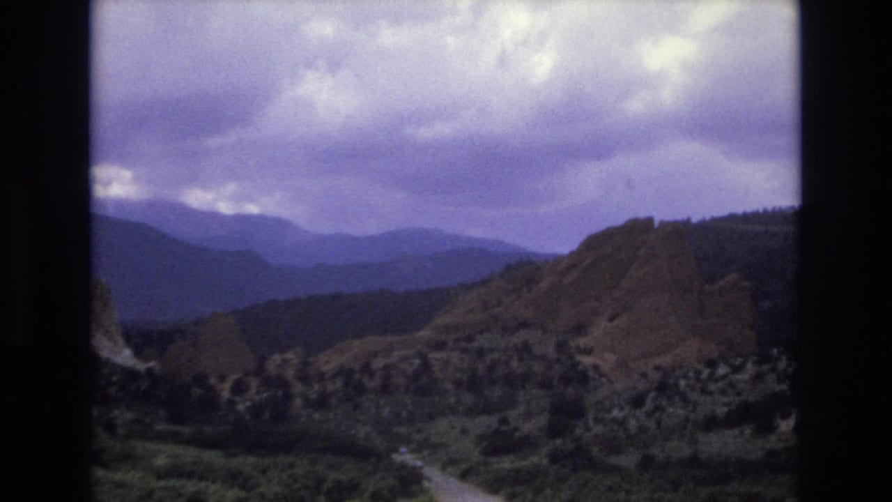 1973 Colorado 8mm frame showing layered mountain ridges, reddish rocky slopes, forested valley terrain, and a cloudy purple-shifted sky.