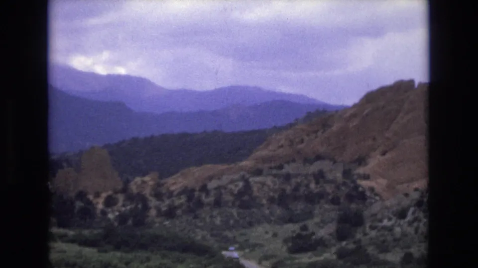 1973 Colorado home-movie still with rocky hillside in the foreground, wooded valley floor, and blue mountain ranges fading into a cloudy sky.