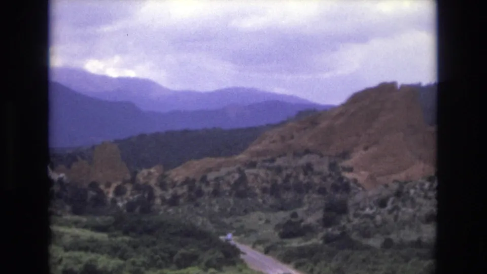 1973 Colorado 8mm frame showing a pale winding road crossing a green forested valley with rocky slopes and distant mountain ridges beyond.