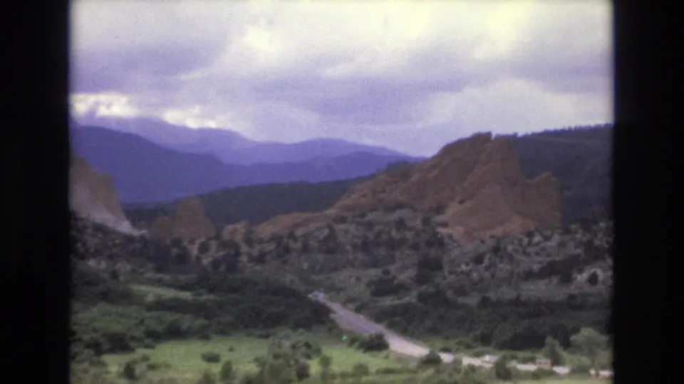 1973 Colorado scenic frame with a winding road, green valley floor, reddish rocky formations, wooded slopes, and layered mountains under an overcast sky.