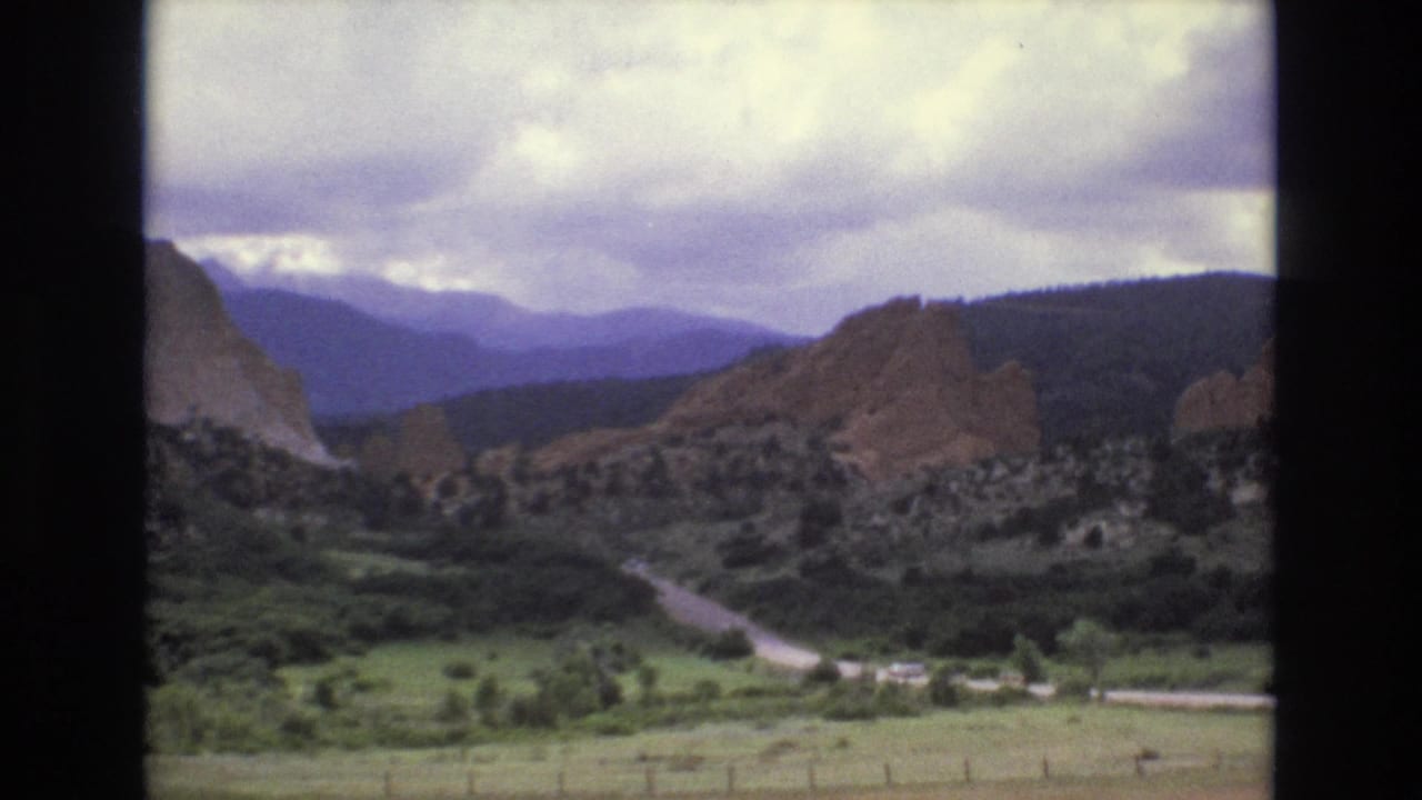 1973 Colorado 8mm frame showing red rock formations, a green valley, a winding road, distant mountains, and a clouded sky.