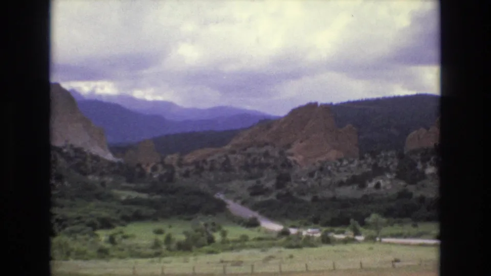 1973 Colorado 8mm frame showing red rock formations, a green valley, a winding road, distant mountains, and a clouded sky.