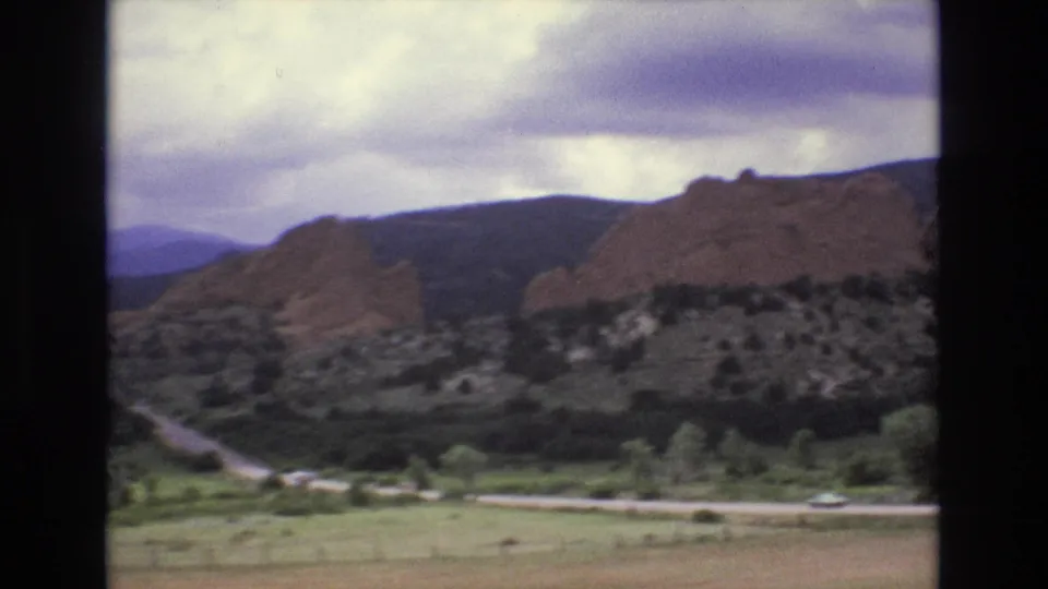 1973 Colorado travel frame with red rock cliffs, foothills, roadside greenery, distant mountains, and overcast sky.