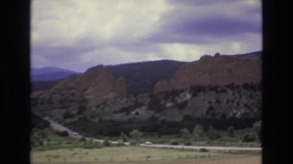 Soft 1973 Colorado 8mm view of red rock formations above a green valley road, with mountains fading into a cloudy background.