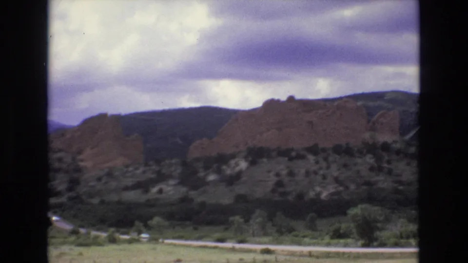 1973 Colorado home-movie frame looking across a green valley road toward red rock ridges and dark mountain slopes under a cloudy sky.