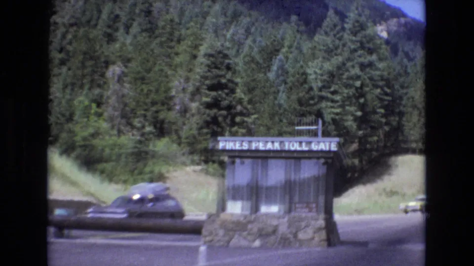 The Pikes Peak Toll Gate booth stands beside a road with period cars passing, backed by evergreen-covered mountain slopes in Colorado archival home-movie footage.