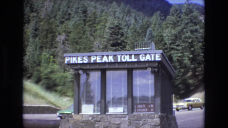 Closer 1970s Colorado view of the Pikes Peak Toll Gate booth, with sign lettering, glass windows, stonework, evergreen trees, and a car on the road behind it.
