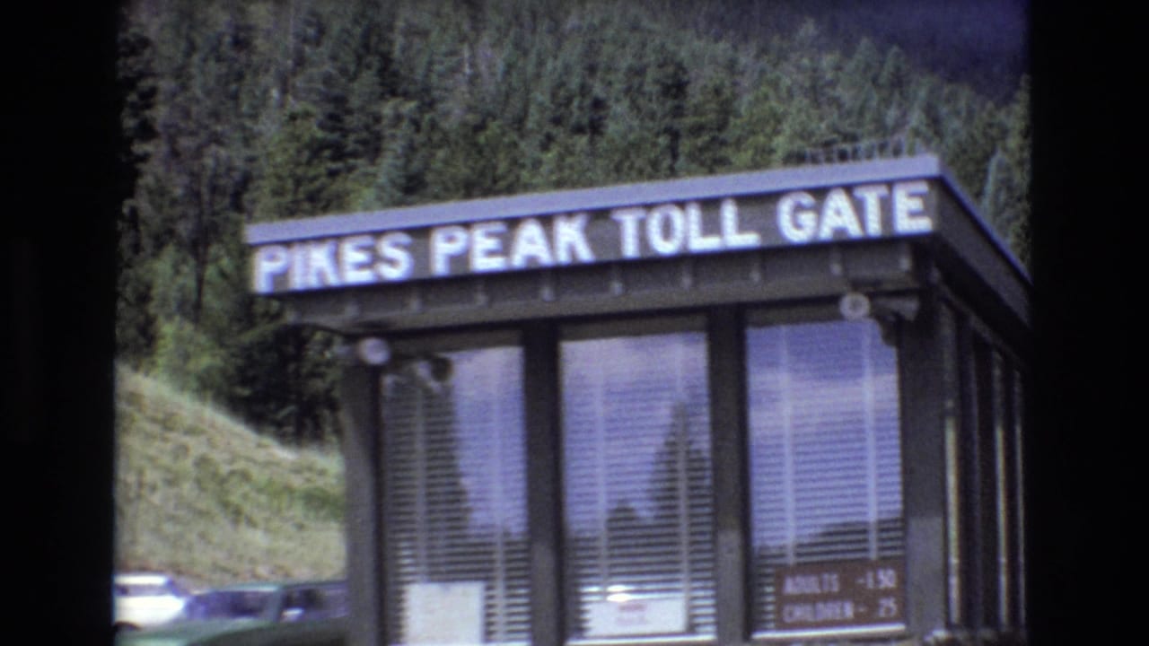 1973 Colorado 8mm frame showing the Pikes Peak Toll Gate booth with readable sign, windows, fee board, parked cars, and evergreen-covered hillside behind it.