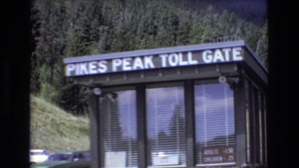 1973 Colorado 8mm frame showing the Pikes Peak Toll Gate booth with readable sign, windows, fee board, parked cars, and evergreen-covered hillside behind it.