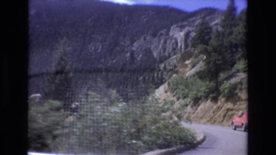 1973 Colorado 8mm frame looking along a winding mountain road bordered by rock cuts, dense evergreens, and a red car entering the curve at the right edge.