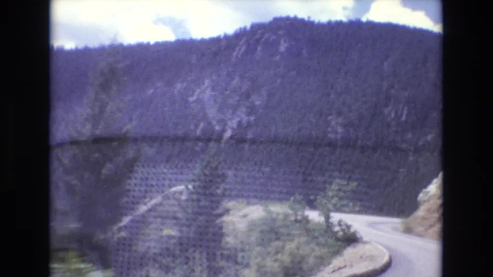 1973 Colorado 8mm frame showing a winding paved road below dark forested hills, exposed rock faces, roadside brush, and soft home-movie motion blur.