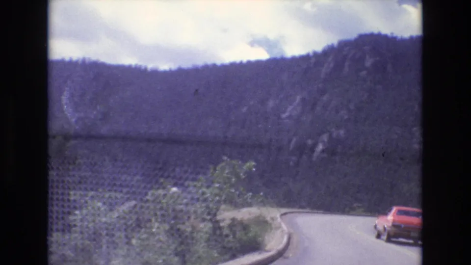 1973 Colorado 8mm frame with a red car rounding a sharp paved curve beside roadside brush, rock walls, and evergreen-covered Colorado mountain slopes.