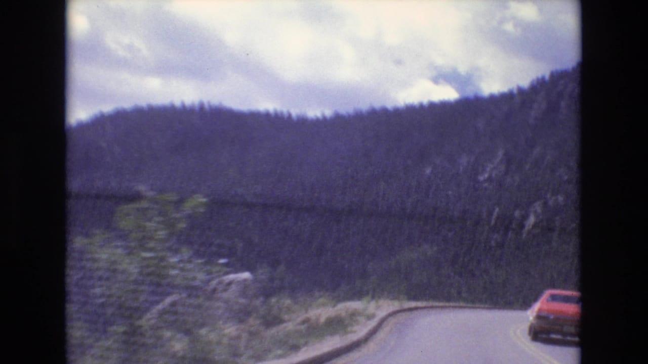 Red car on a curving mountain road below forested hills and cloudy sky.