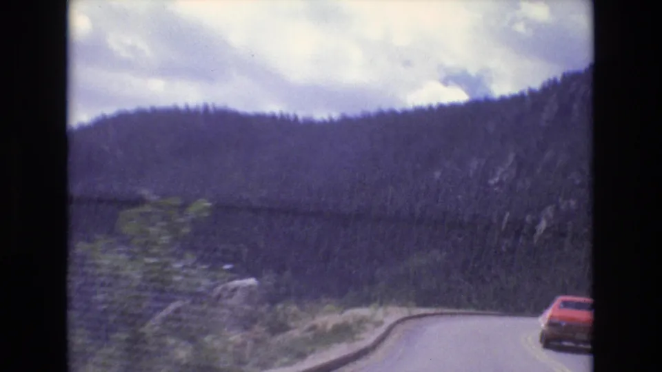 1973 Colorado 8mm frame showing a red car on a winding asphalt road beside forested mountain slopes under a partly cloudy sky.